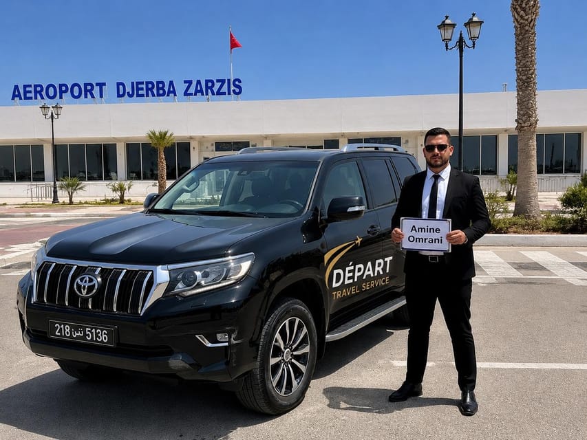 A driver in a black suit holding a name sign beside a black SUV in front of the Djerba-Zarzis airport terminal