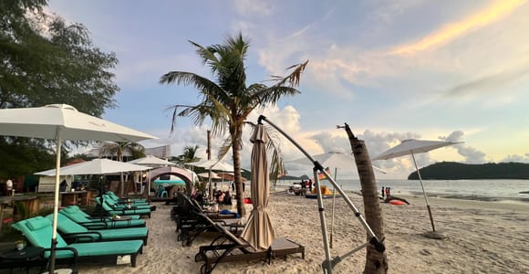 Sun loungers and white parasols on pale sand at dusk, palms leaning over a beach bar, a wooded headland across the bay under an orange sky