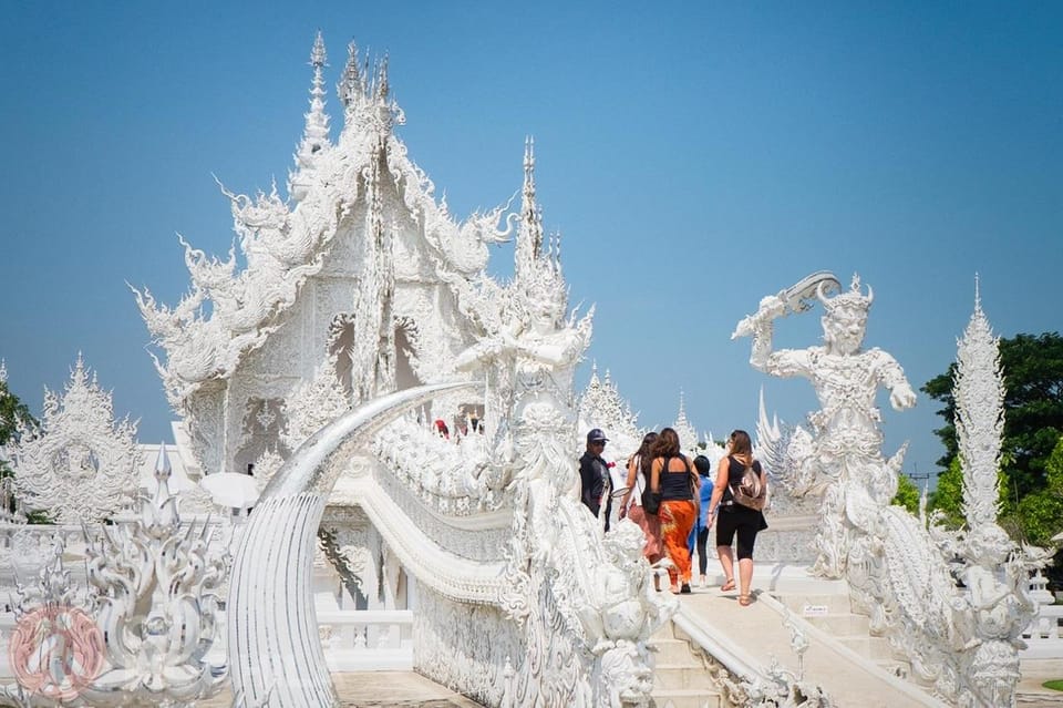 Visitors crossing the ornate white bridge towards the mirrored facade of Wat Rong Khun under a cloudless sky