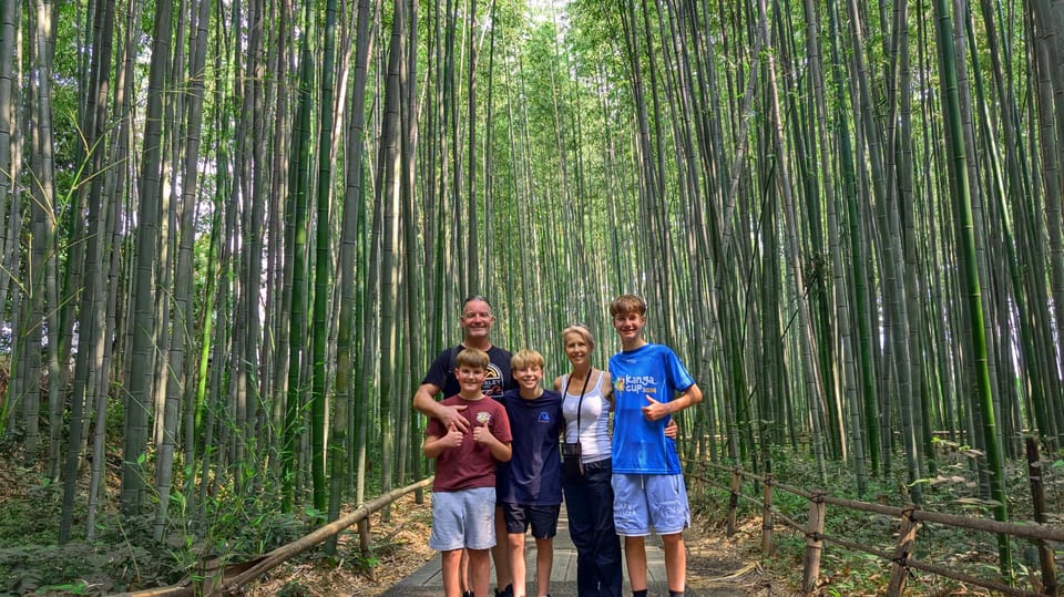 A family of five standing together on a paved path through a dense bamboo grove, low wooden railings on both sides, green light overhead