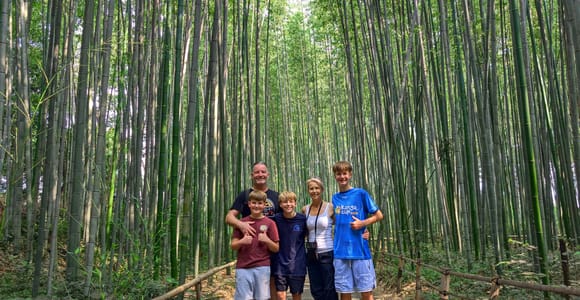 A family of five standing together on a paved path through a dense bamboo grove, low wooden railings on both sides, green light overhead