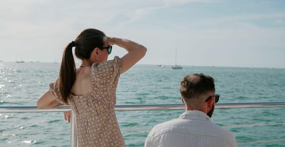 Two passengers at the rail of an excursion boat, seen from behind, looking out over calm green-blue water with a single sailboat on the horizon