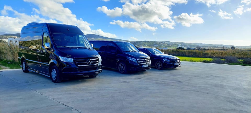 Three black vans and saloon cars parked on a paved forecourt beside green fields and low hills
